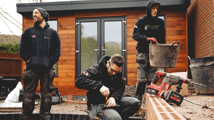 four tradies working outside a wooden panel house with power tools and dirty buckets