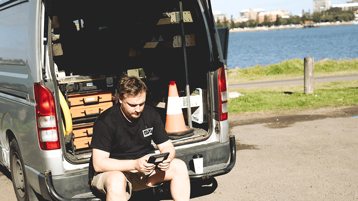 tradie sitting at the boot of company van looking at cellphone