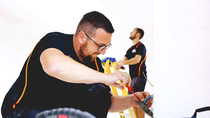 tradie in the forefront working on power switched attached to wall and another tradie at the back on a ladder looking at the ceiling